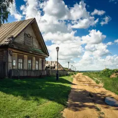 Drewniany dom i polna droga, której szerokość dojazdowa jest widoczna, prowadzą przez wieś pod błękitnym niebem z chmurami.