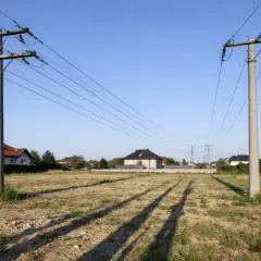 Linia średniego napięcia biegnie nad niezabudowaną działką, z domami w oddali. Widać wyraźną odległość budynku od linii średniego napięcia.