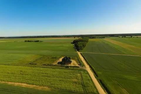 Dom w budowie na tle pól i lasu, co wymaga pozwolenia na budowę. W oddali widać farmę wiatrową.